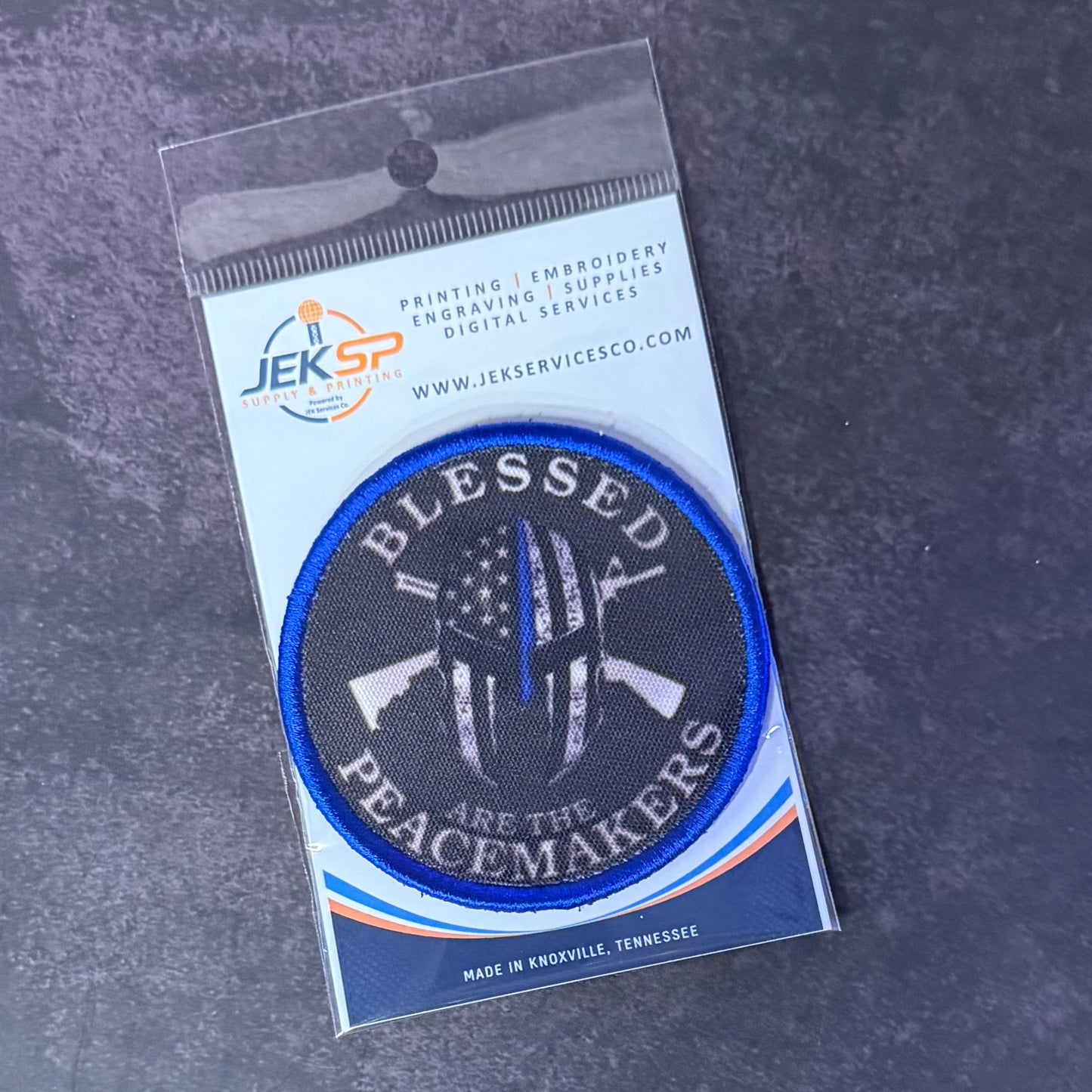 An embroidered circular patch in its original clear plastic packaging, displayed on a dark textured gray background. The patch features a black background with a blue border, white text reading 'BLESSED PEACEMAKERS', and a central white peace symbol with a thin red line and an American flag pattern.