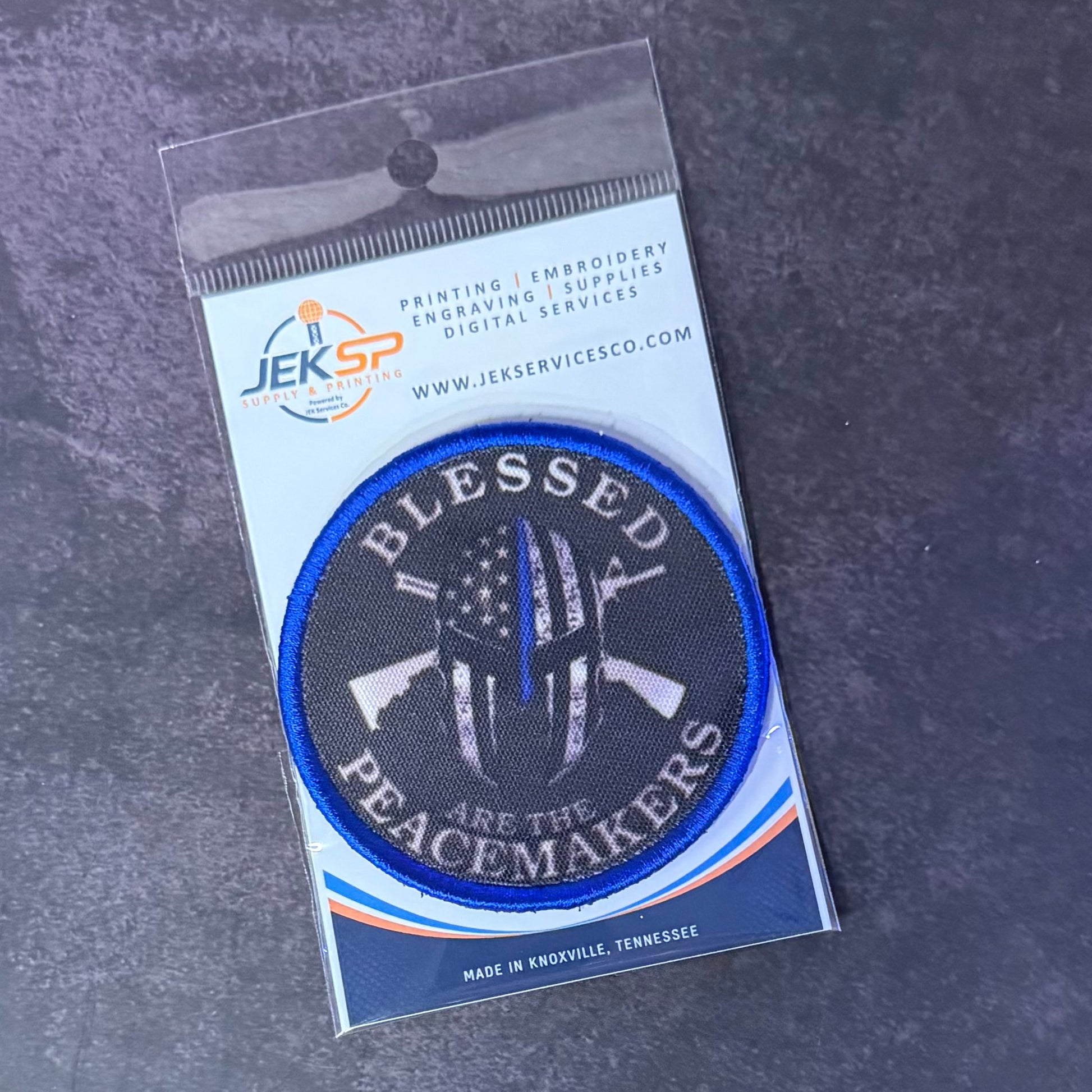 An embroidered circular patch in its original clear plastic packaging, displayed on a dark textured gray background. The patch features a black background with a blue border, white text reading 'BLESSED PEACEMAKERS', and a central white peace symbol with a thin red line and an American flag pattern.