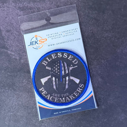 An embroidered circular patch in its original clear plastic packaging, displayed on a dark textured gray background. The patch features a black background with a blue border, white text reading 'BLESSED PEACEMAKERS', and a central white peace symbol with a thin red line and an American flag pattern.