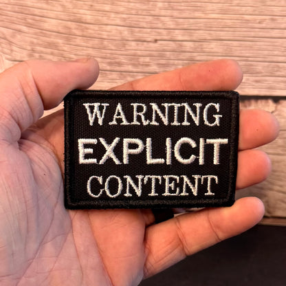 A rectangular black embroidered patch with white text reading 'WARNING EXPLICIT CONTENT' held in a person's hand against a wood plank background.