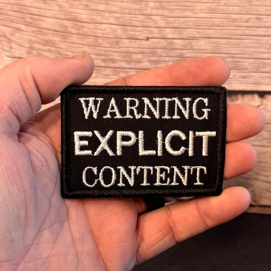 A rectangular black embroidered patch with white text reading 'WARNING EXPLICIT CONTENT' held in a person's hand against a wood plank background.