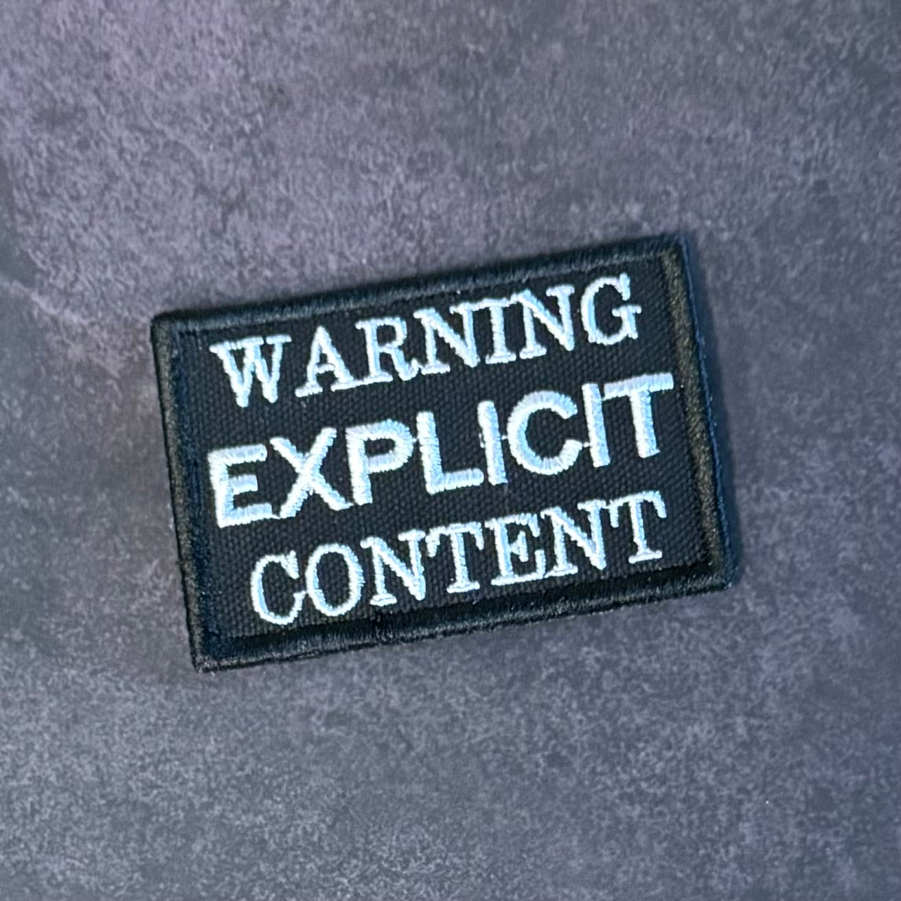 A rectangular black embroidered patch with white text reading 'WARNING EXPLICIT CONTENT' on a textured gray background.