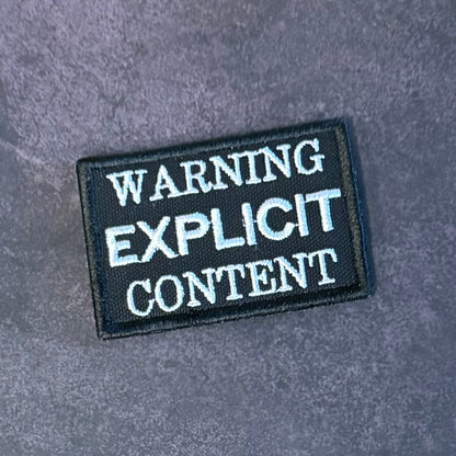 A rectangular black embroidered patch with white text reading 'WARNING EXPLICIT CONTENT' on a textured gray background.