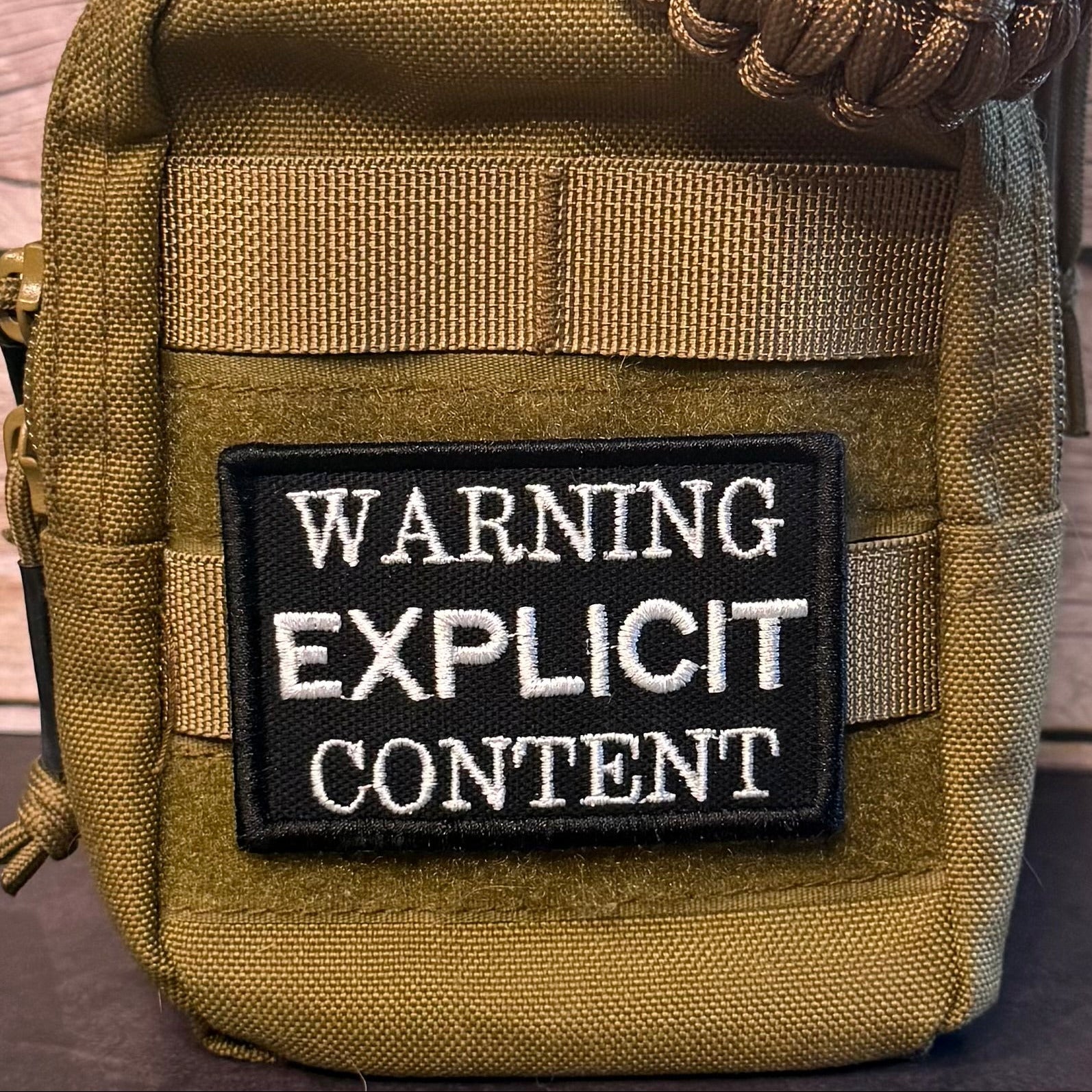 Close-up of a tactical pouch in olive drab and tan webbing, featuring a black rectangular patch with white text that reads 'WARNING EXPLICIT CONTENT'.