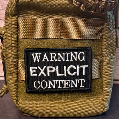 Close-up of a tactical pouch in olive drab and tan webbing, featuring a black rectangular patch with white text that reads 'WARNING EXPLICIT CONTENT'.