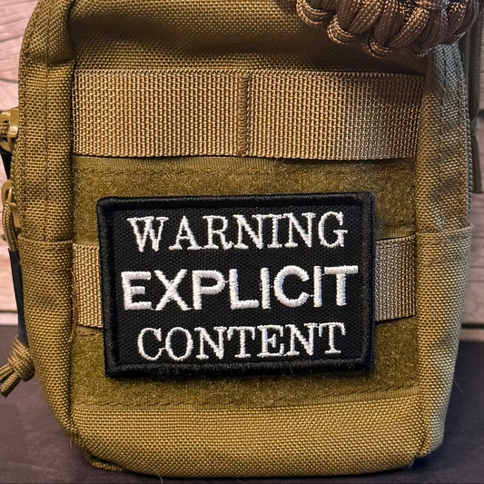 Close-up of a tactical pouch in olive drab and tan webbing, featuring a black rectangular patch with white text that reads 'WARNING EXPLICIT CONTENT'.