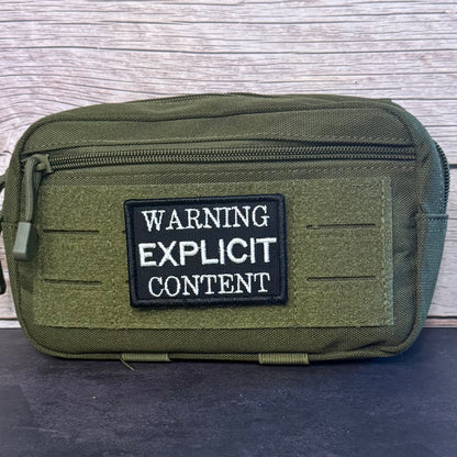 An olive green tactical pouch with a black and white 'WARNING EXPLICIT CONTENT' patch on the front, set against a light wood panel background.