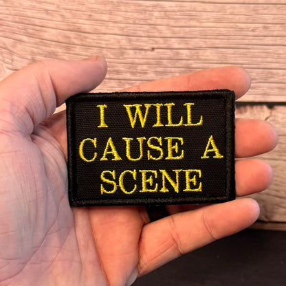 A hand holding a rectangular black embroidered patch with yellow text that reads 'I WILL CAUSE A SCENE' against a wood-grain background.
