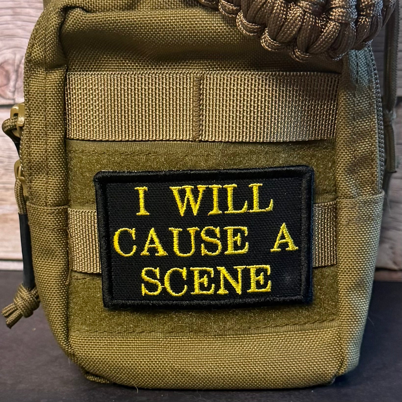 A close-up of a tactical pouch in olive drab green nylon with a black rectangular patch featuring yellow embroidered text that reads 'I WILL CAUSE A SCENE'.