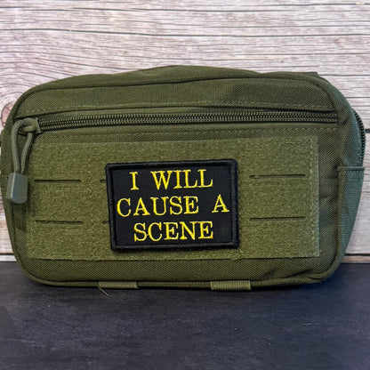 An olive green tactical pouch with a black rectangular patch featuring yellow embroidered text that reads 'I WILL CAUSE A SCENE'. The pouch is shown on a dark surface with a light wood background.