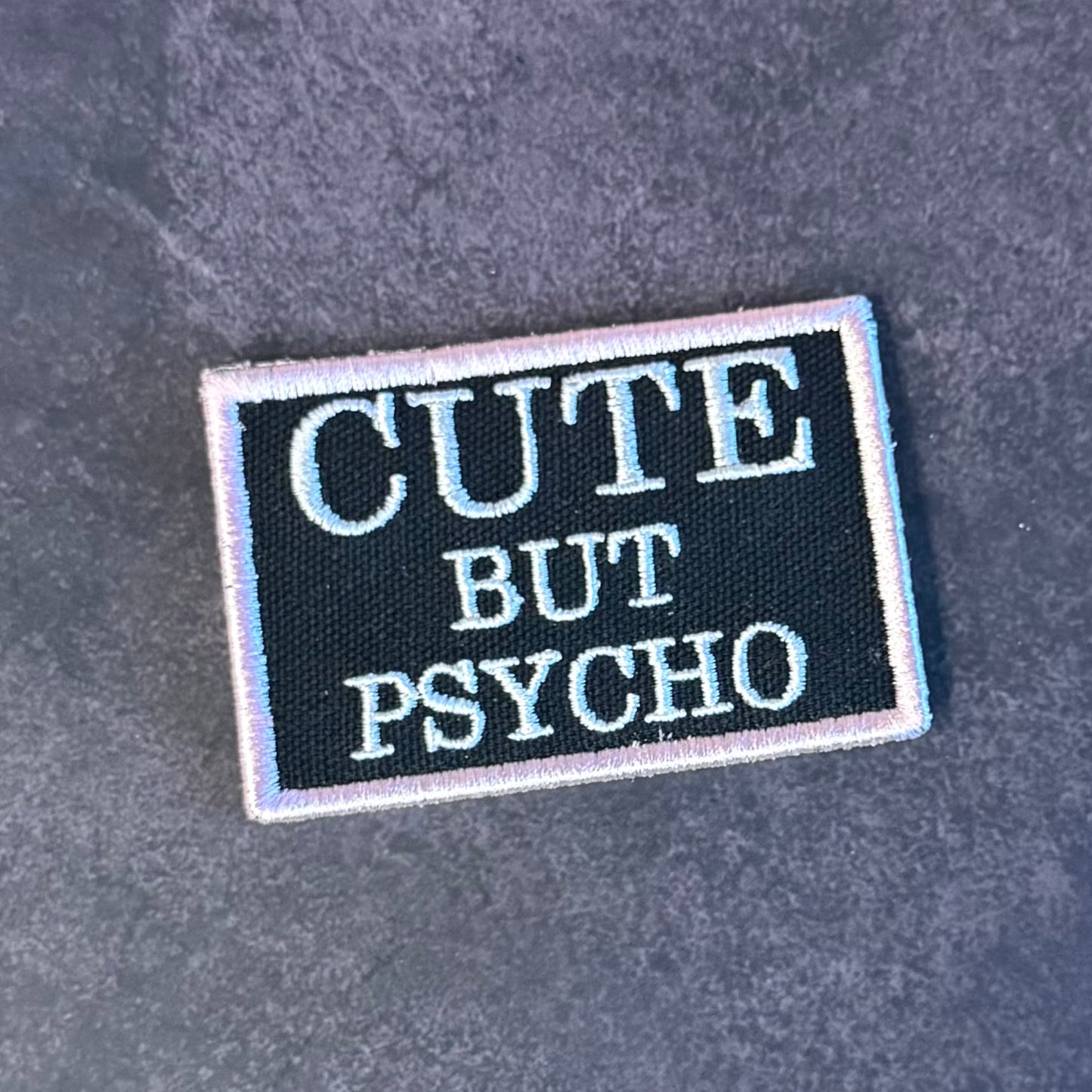 A rectangular embroidered patch with the text 'CUTE BUT PSYCHO' in white lettering on a black background, finished with a white border, shown on a dark gray textured fabric background.
