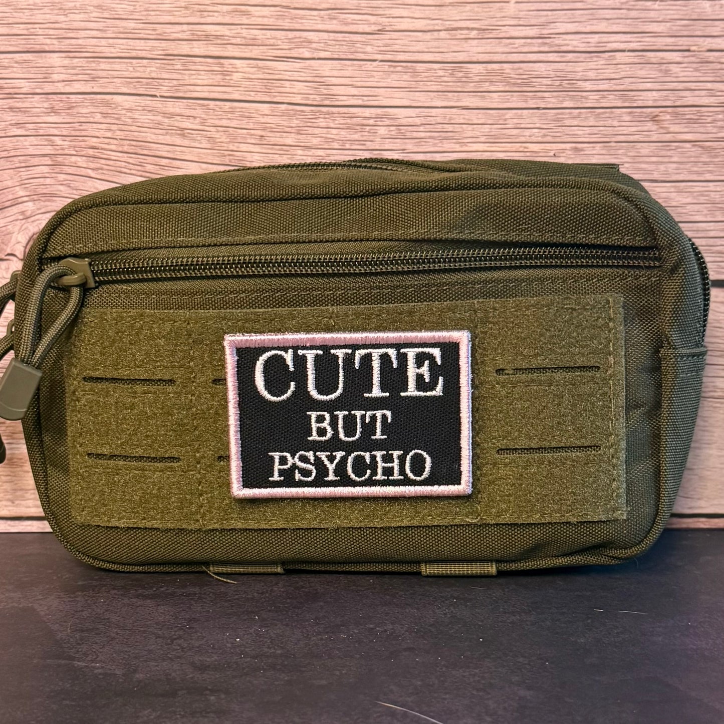 An olive green tactical pouch with a black rectangular patch that reads 'CUTE BUT PSYCHO' in white text, set against a wood-grain background.