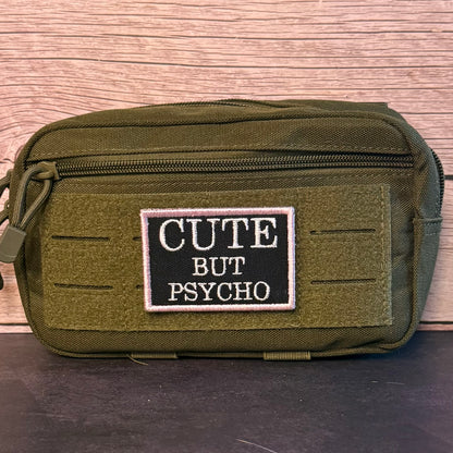 An olive green tactical pouch with a black rectangular patch that reads 'CUTE BUT PSYCHO' in white text, set against a wood-grain background.