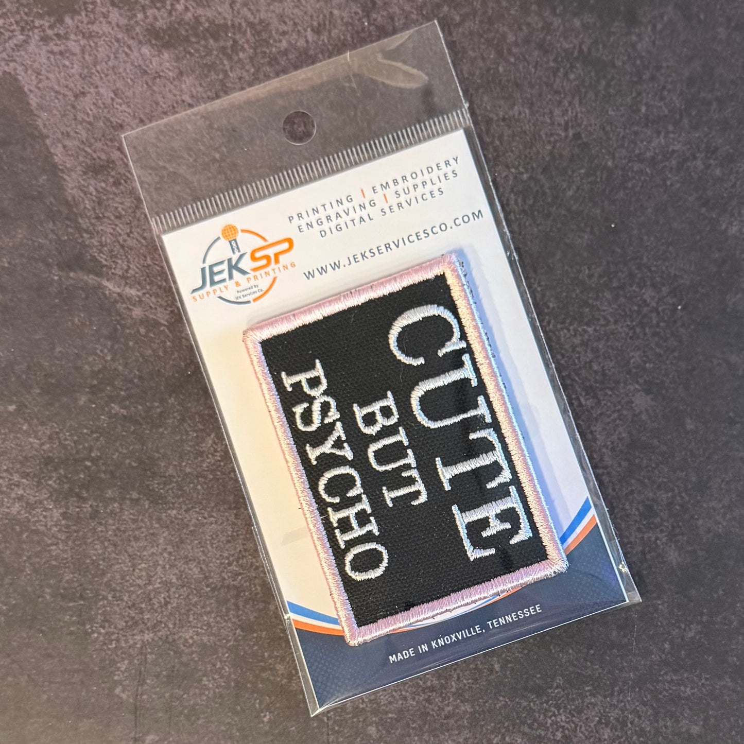 A packaged embroidered patch on a clear plastic sleeve over a gray textured surface. The patch is black with white text reading 'CUTE BUT PSYCHO' and a thin pink border.