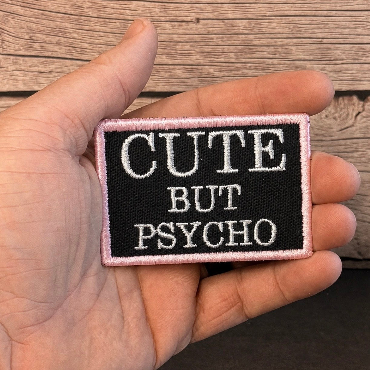 A rectangular embroidered patch held in a hand, featuring the text 'CUTE BUT PSYCHO' in white embroidery on a black background with a light pink border, set against a rustic wooden background.