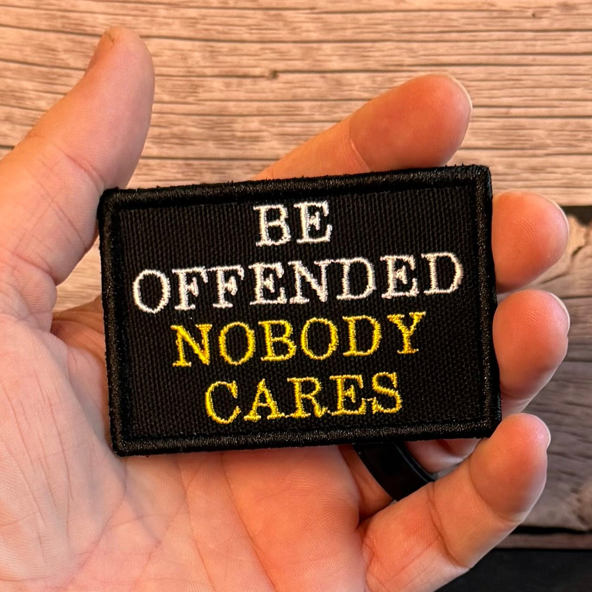 A hand holding a rectangular black embroidered patch with the text 'BE OFFENDED NOBODY CARES' in white and yellow lettering, set against a rustic wooden background.