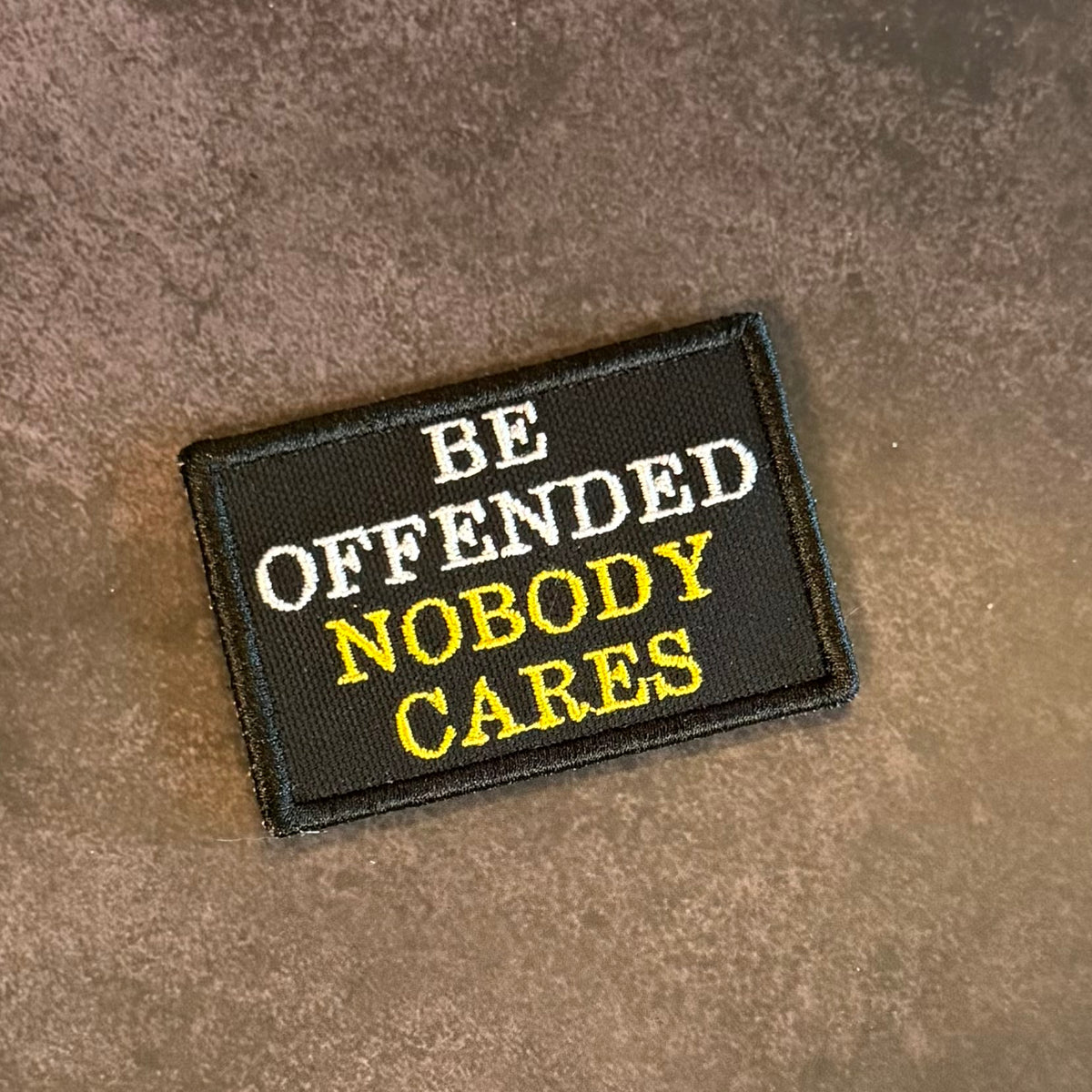 A rectangular embroidered patch with the text 'BE OFFENDED NOBODY CARES' in white and yellow lettering on a black background, placed on a textured brown surface.