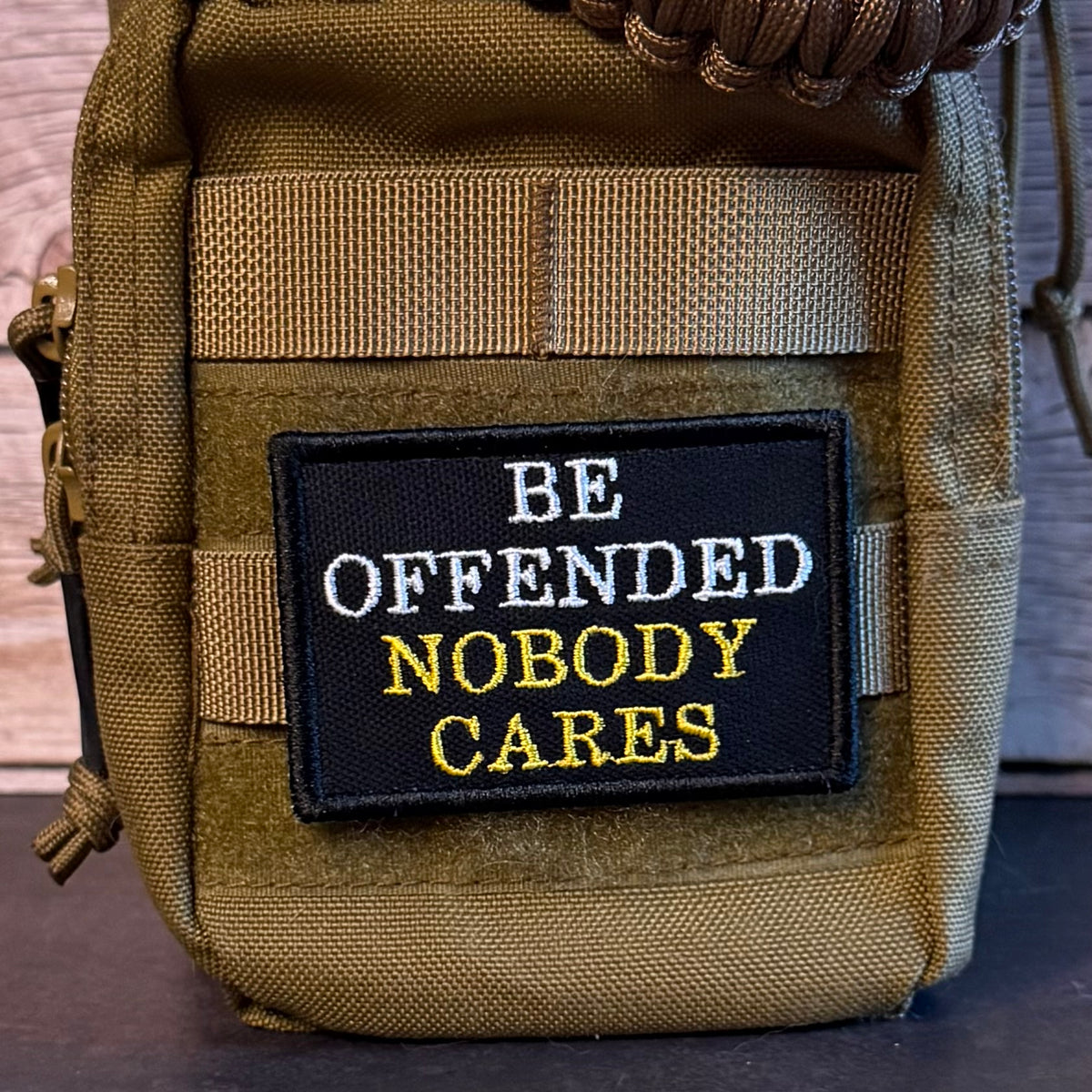 A close-up of a tactical pouch in olive drab and coyote brown webbing, featuring a black rectangular embroidered patch with the text 'BE OFFENDED NOBODY CARES' in white and yellow.