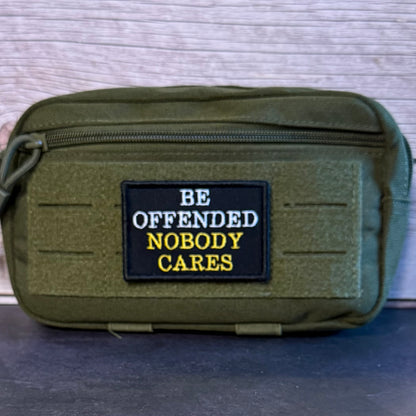 An olive green tactical pouch with a black embroidered patch that reads 'BE OFFENDED NOBODY CARES' in white and yellow text, set against a light wood background.