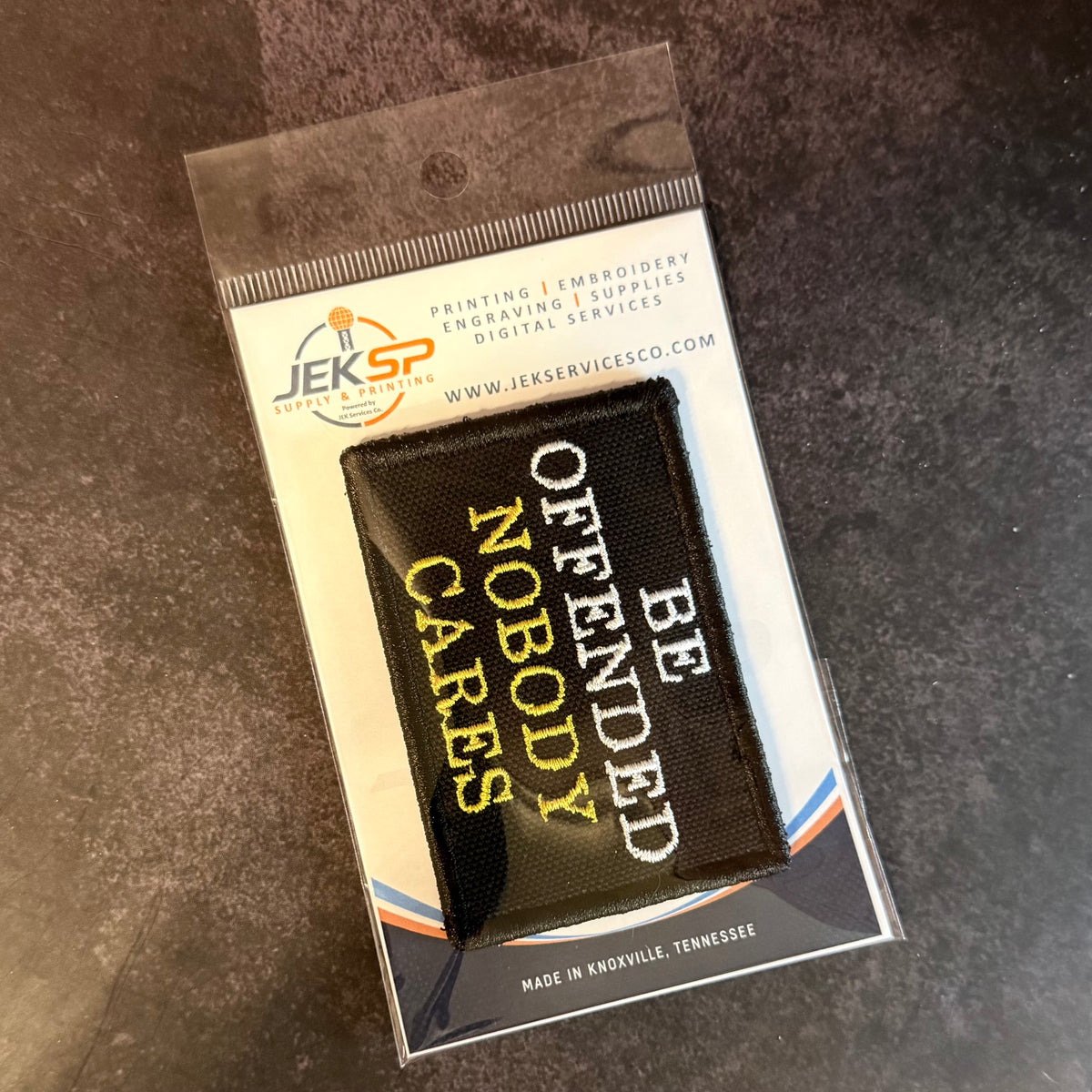 A black embroidered patch with white and yellow text reading 'BE OFFENDED NOBODY NOBODY CARES' in a clear plastic sleeve with a branded JEKSP card insert, photographed on a dark textured surface.