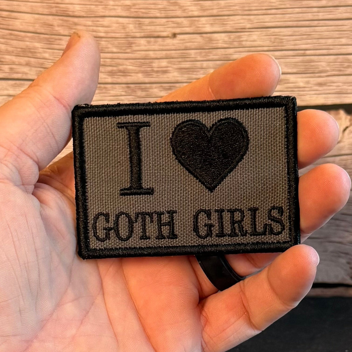 A hand holding a rectangular embroidered patch that reads 'I [heart] GOTH GIRLS' in black on a gray fabric background, with a wood plank surface behind it.