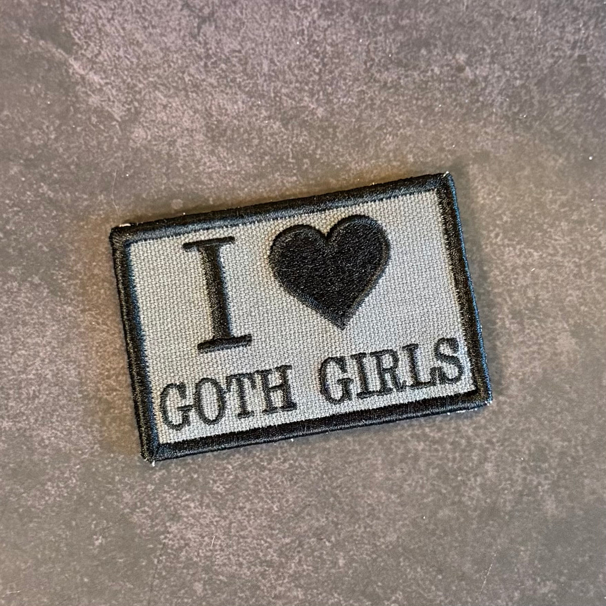 An embroidered rectangular patch with the text 'I' followed by a black heart and 'GOTH GIRLS' in black lettering on a light gray fabric background, finished with a black stitched border, shown on a textured gray surface.