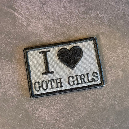 An embroidered rectangular patch with the text 'I' followed by a black heart and 'GOTH GIRLS' in black lettering on a light gray fabric background, finished with a black stitched border, shown on a textured gray surface.