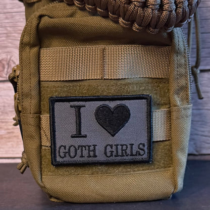 A close-up of a tactical pouch in olive drab and coyote brown webbing, featuring a rectangular gray patch with black embroidery that reads 'I ♥ GOTH GIRLS'.