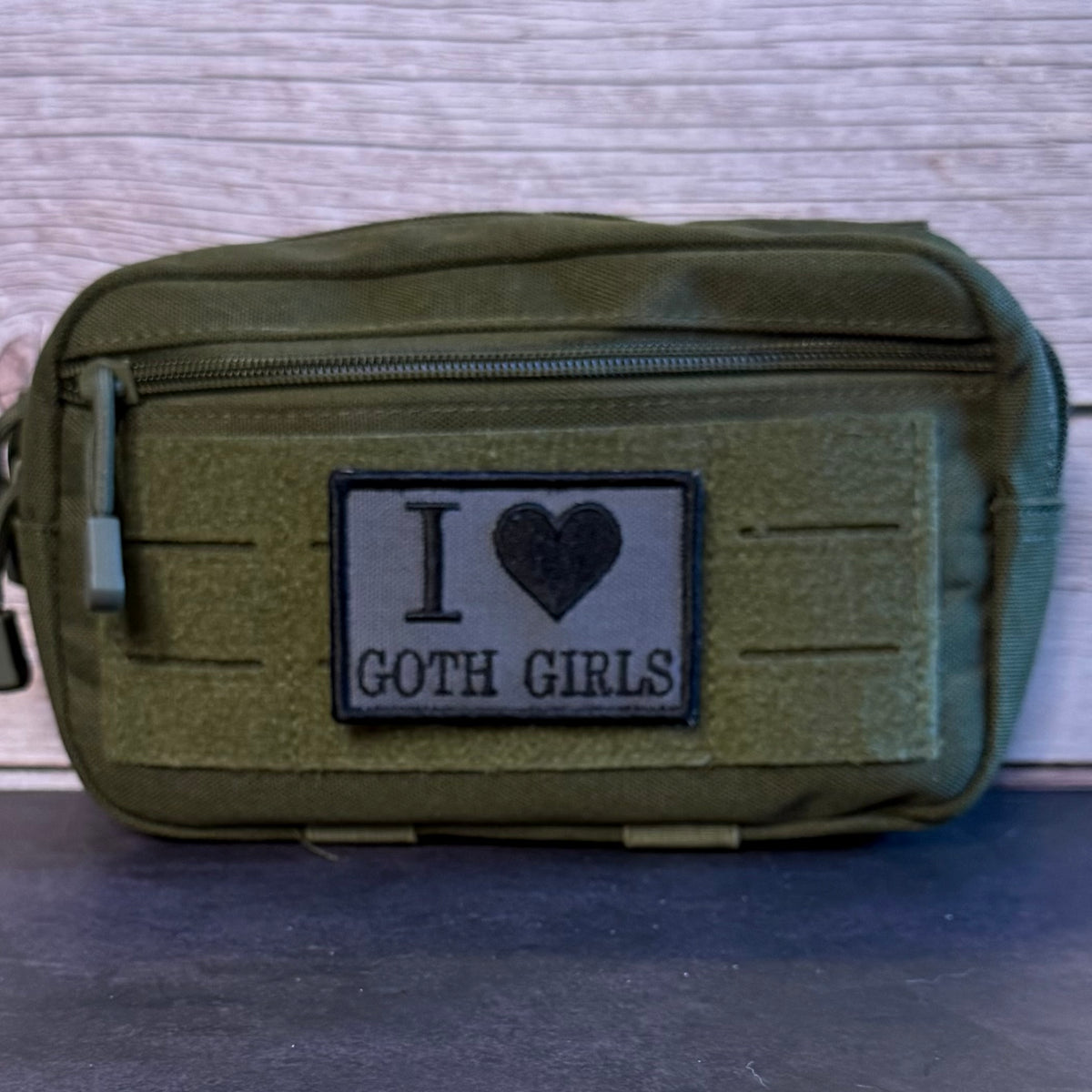 An olive green tactical pouch with a front zipper and a black rectangular patch that reads 'I [heart] GOTH GIRLS' in black text, shown against a light wood background.