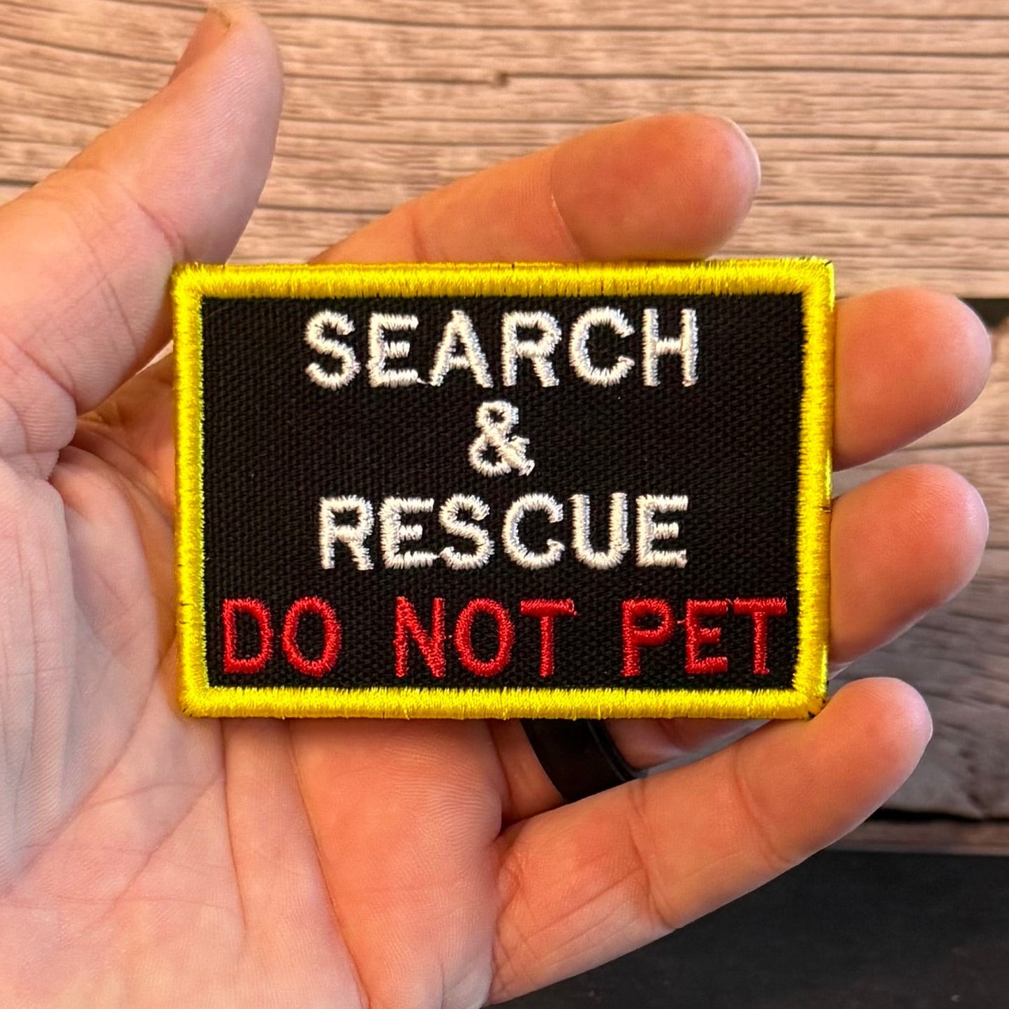 A hand holding a rectangular embroidered patch that reads 'SEARCH & RESCUE DO NOT PET' in white and red text on a black background with a yellow border, set against a wooden surface.