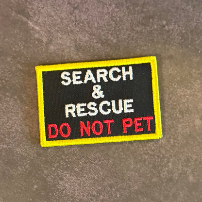 An embroidered rectangular patch with a black center, yellow border, and text reading 'SEARCH & RESCUE DO NOT PET' in white and red, placed on a textured gray surface.