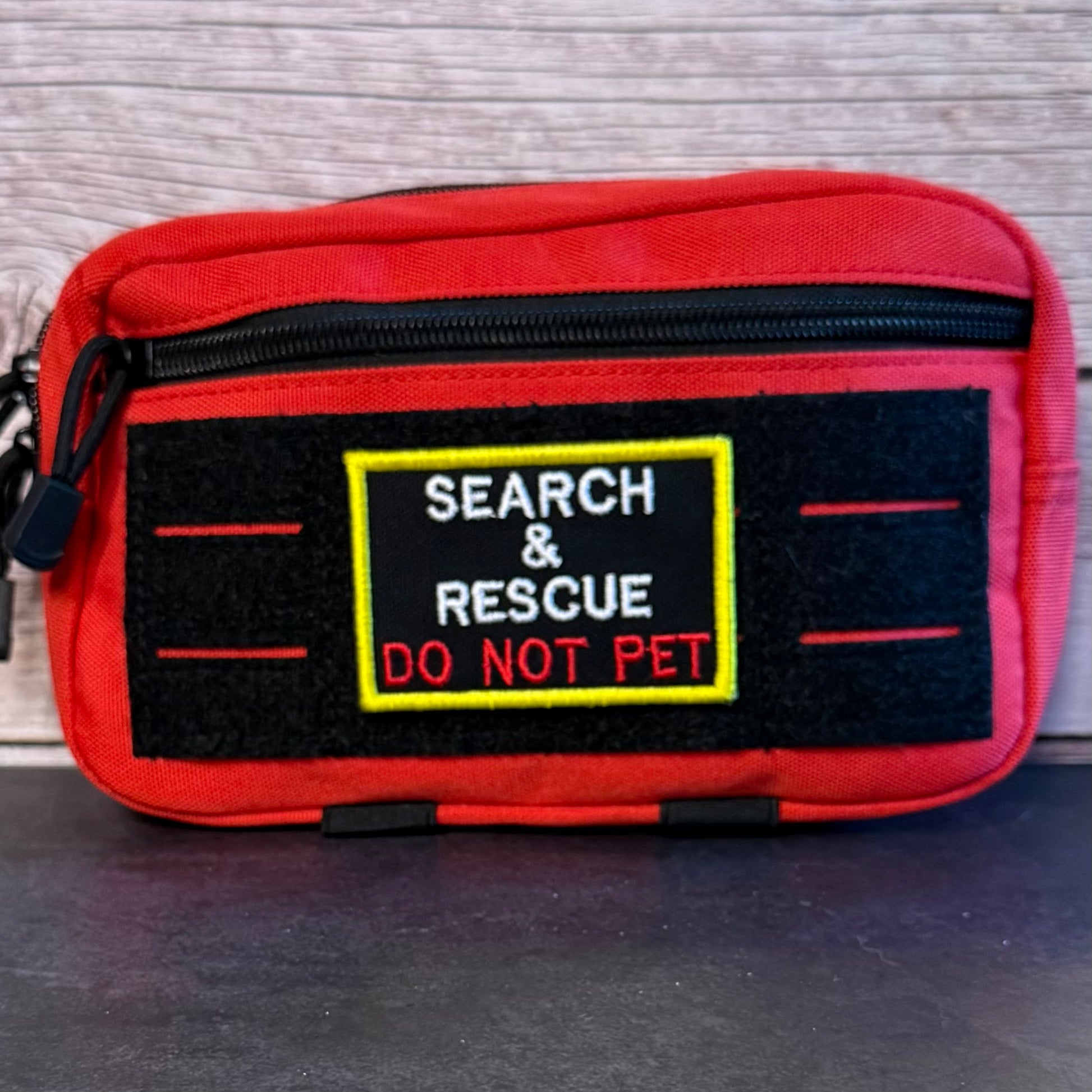 A red zippered pouch with a black front patch that reads 'SEARCH & RESCUE DO NOT PET' in white and red text, set against a wood-grain background.