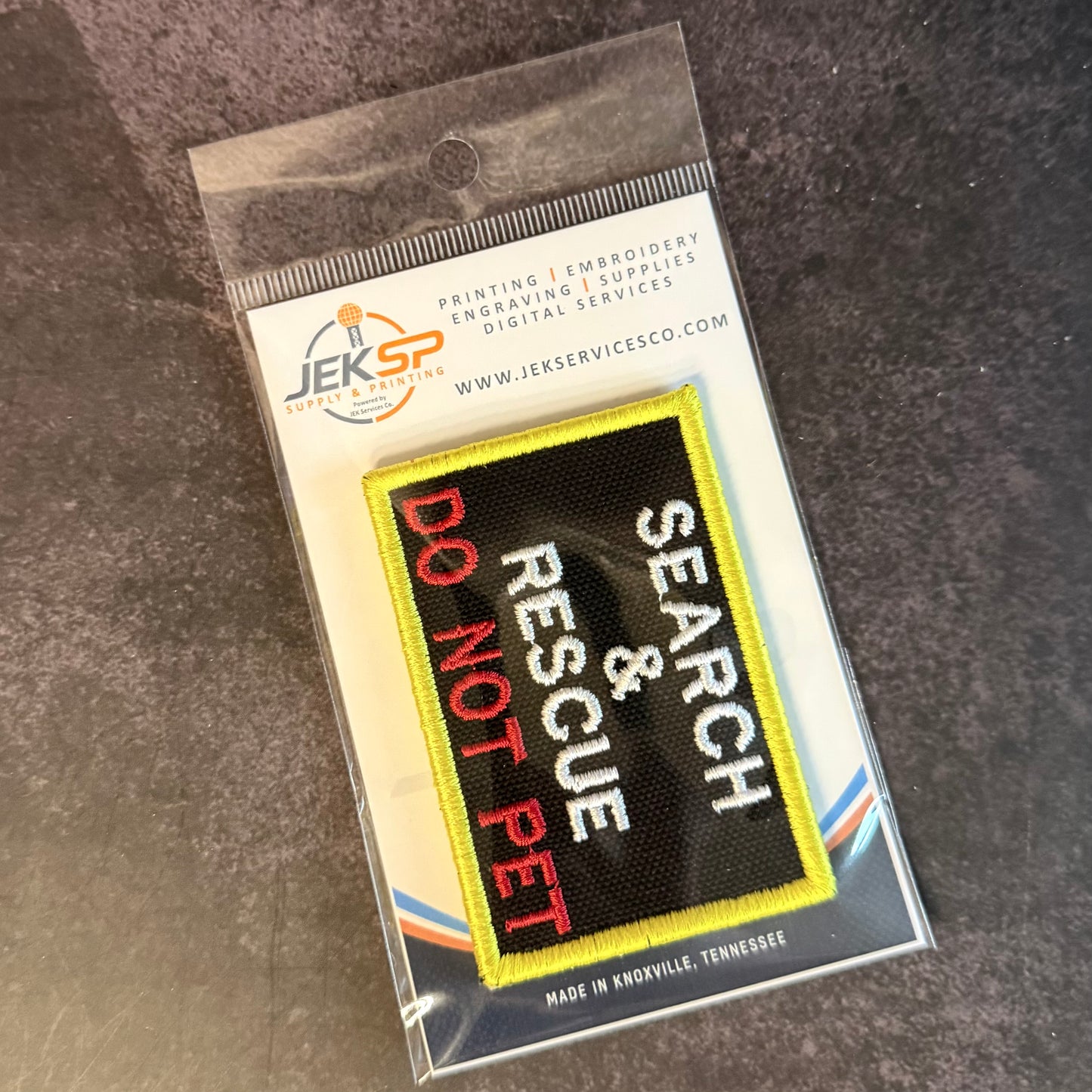 An embroidered patch in clear plastic packaging, featuring a black background with yellow border, white text reading 'SEARCH & RESCUE', and red text reading 'DO NOT PET'. The packaging header shows the JEK SP brand.