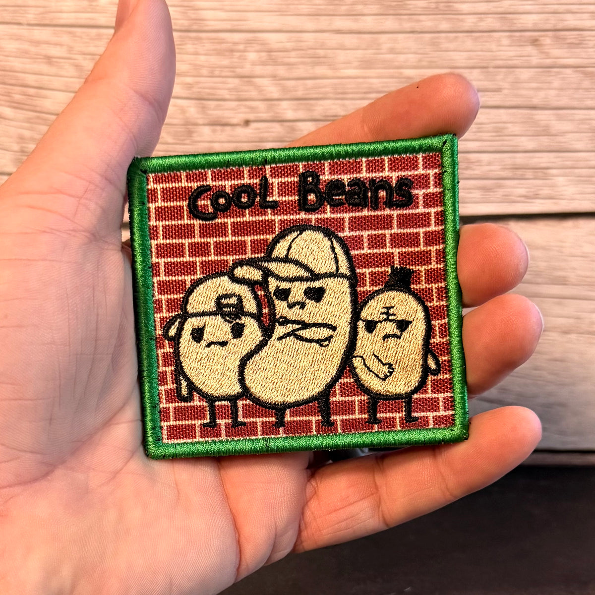 A hand holding a square embroidered patch featuring three cartoon beans on a red brick background with the text 'Cool Beans' and a green border.