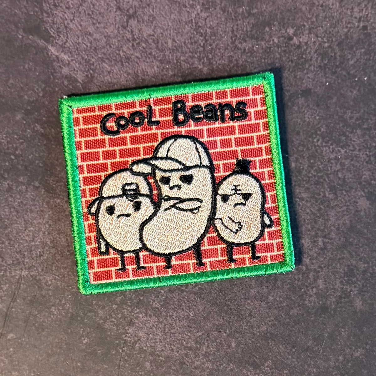A square embroidered patch featuring three cartoon beans wearing caps against a red brick pattern background, with the text 'Cool Beans' at the top and a green stitched border, shown on a dark textured surface.