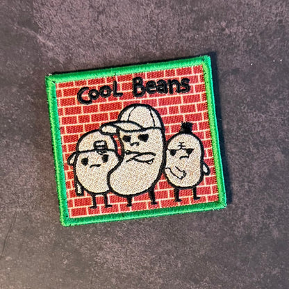 A square embroidered patch featuring three cartoon beans wearing caps against a red brick pattern background, with the text 'Cool Beans' at the top and a green stitched border, shown on a dark textured surface.