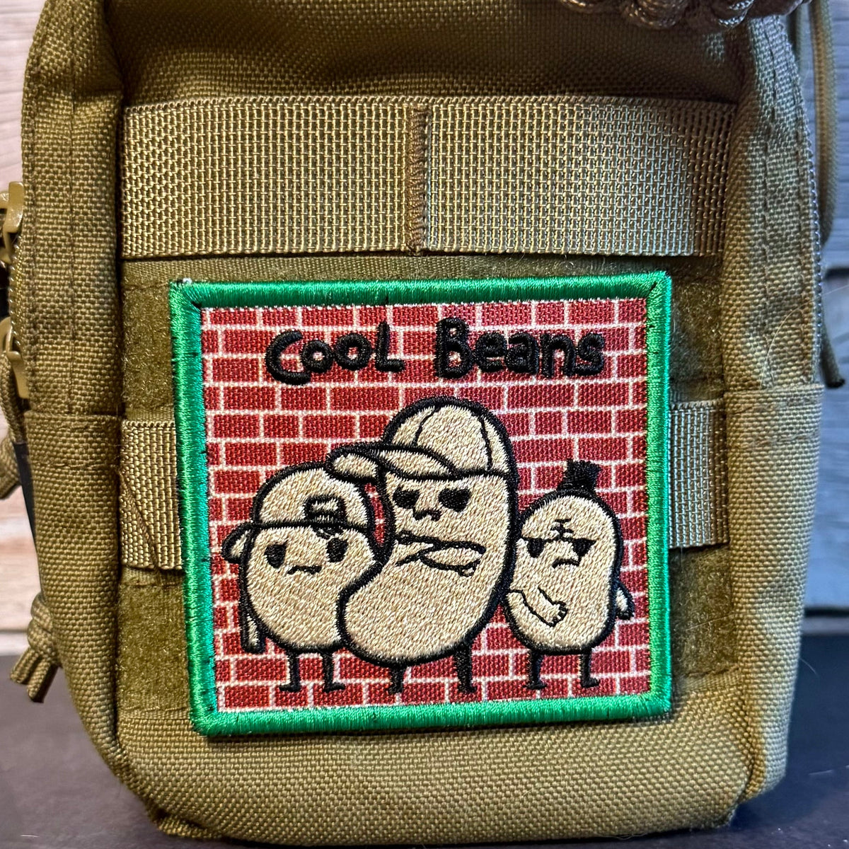 An embroidered patch featuring three cartoon beans on a red brick wall with the text 'Cool Beans' and a green border, attached to an olive drab tactical pouch.
