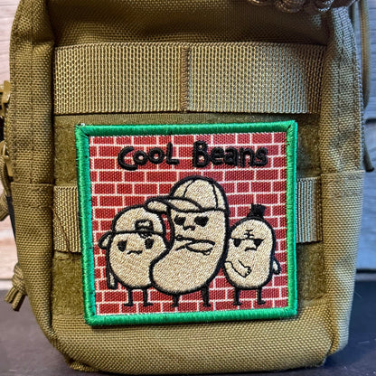 An embroidered patch featuring three cartoon beans on a red brick wall with the text 'Cool Beans' and a green border, attached to an olive drab tactical pouch.