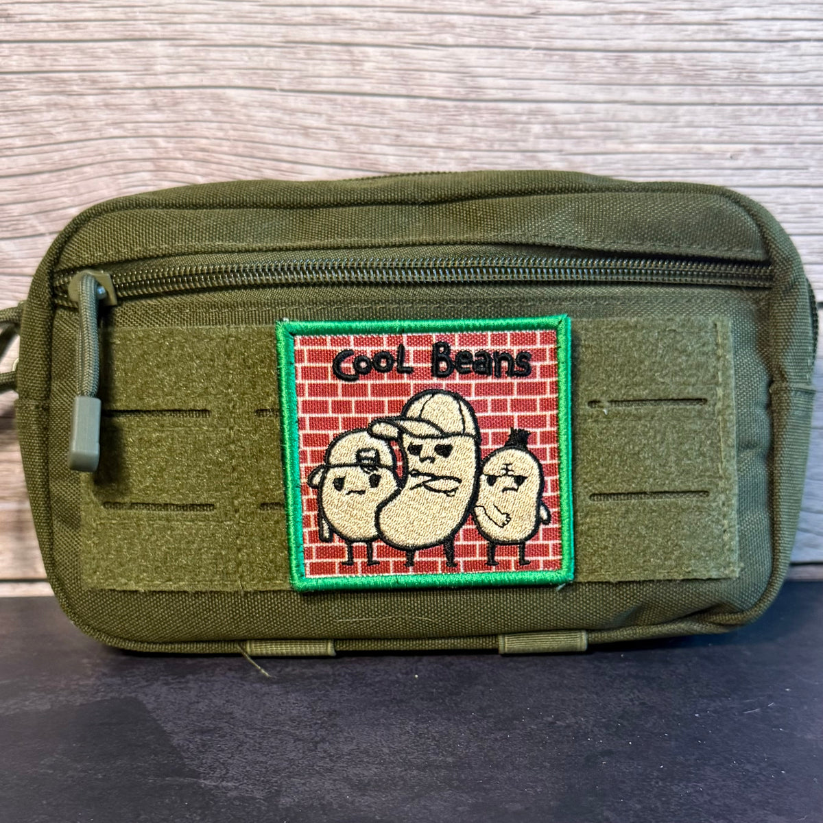 An olive green tactical pouch with a front zipper and a rectangular embroidered patch featuring three cartoon beans on a red brick background with the text 'Cool Beans'.