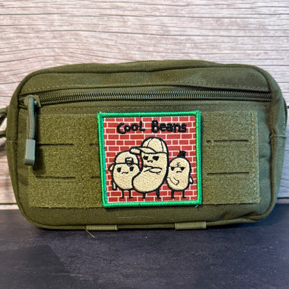 An olive green tactical pouch with a front zipper and a rectangular embroidered patch featuring three cartoon beans on a red brick background with the text 'Cool Beans'.