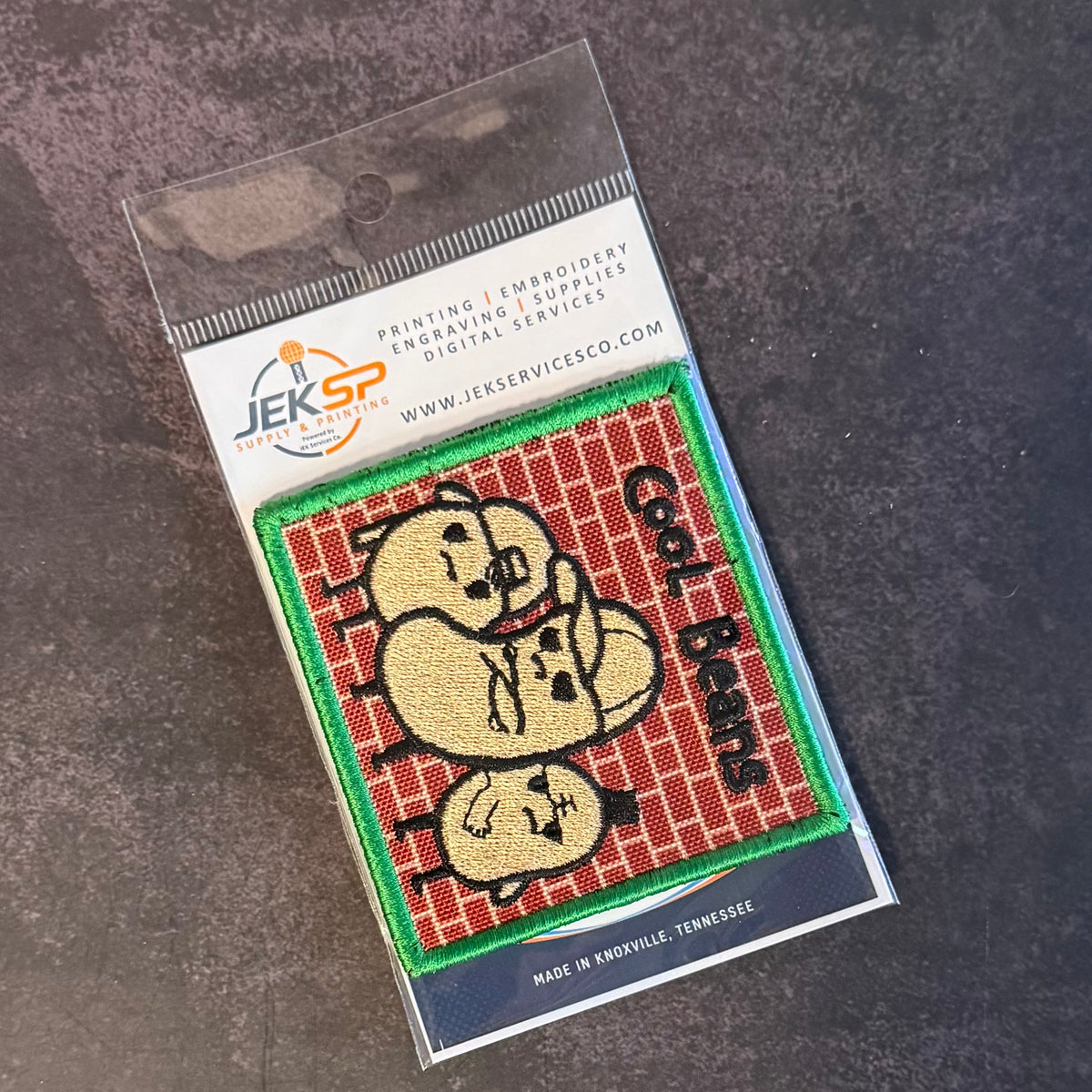 An embroidered patch in its clear plastic packaging, featuring three cartoonish beige heads on a red brick background with green borders and black text reading 'Call of Thieves'. The packaging header shows the JEKSP brand logo and contact details.