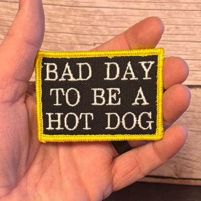 A hand holding a rectangular embroidered patch with the text 'BAD DAY TO BE A HOT DOG' in white on a black background, bordered in bright yellow, against a wood plank background.