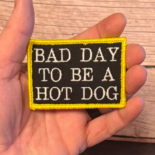 A hand holding a rectangular embroidered patch with the text 'BAD DAY TO BE A HOT DOG' in white on a black background, bordered in bright yellow, against a wood plank background.