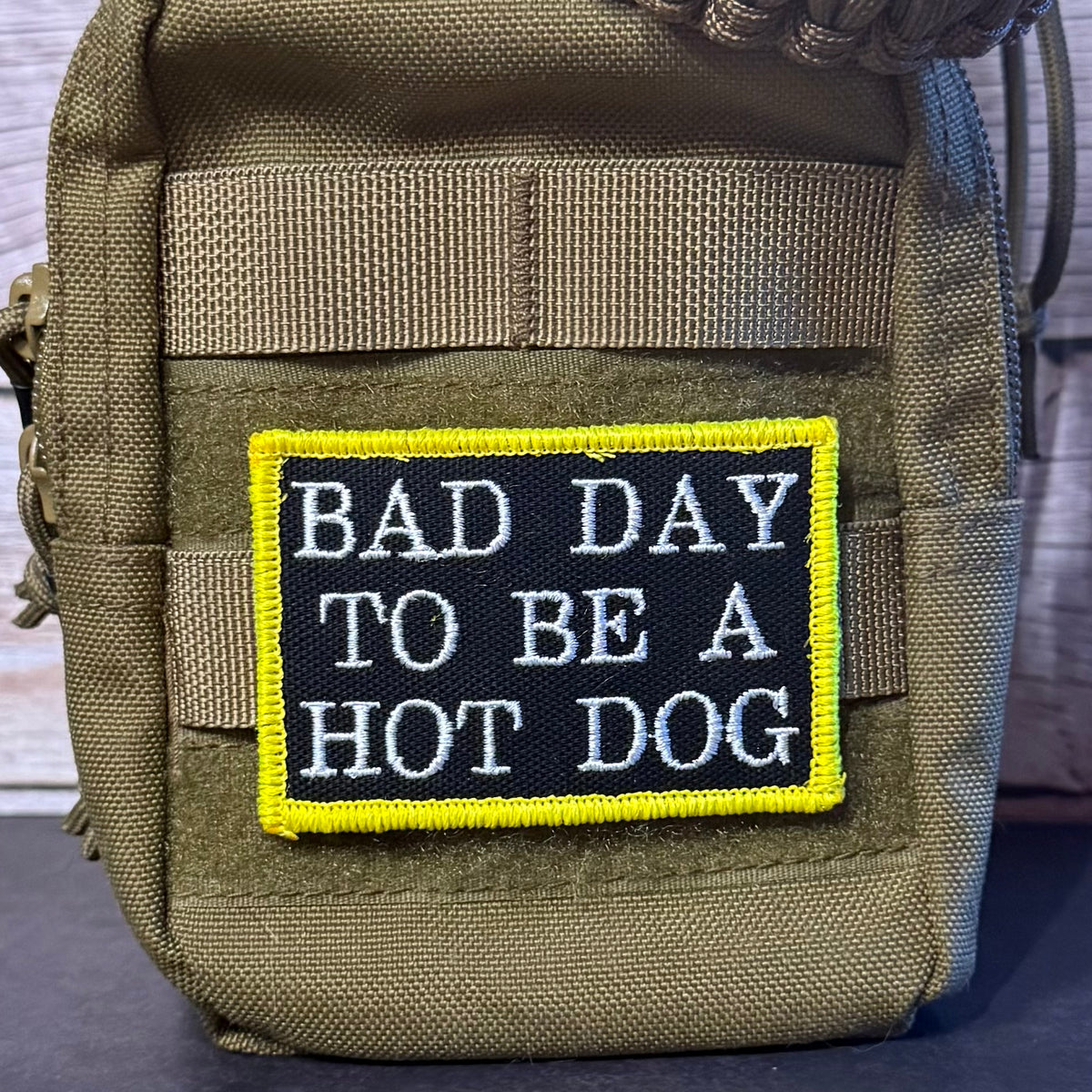 A tactical pouch in olive drab green with a rectangular embroidered patch that reads 'BAD DAY TO BE A HOT DOG' in white text on a black background with a bright yellow border.