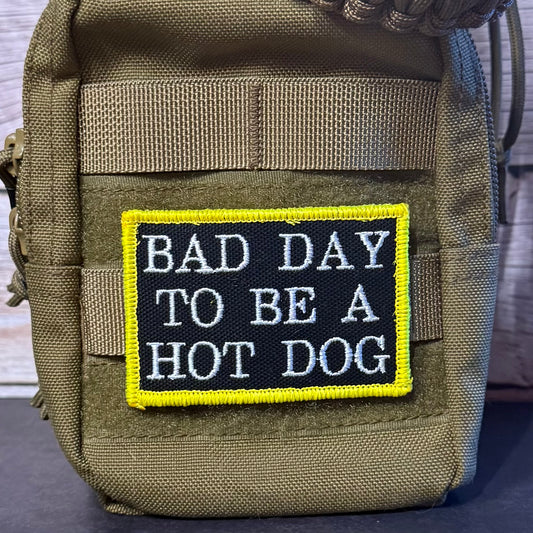 A tactical pouch in olive drab green with a rectangular embroidered patch that reads 'BAD DAY TO BE A HOT DOG' in white text on a black background with a bright yellow border.
