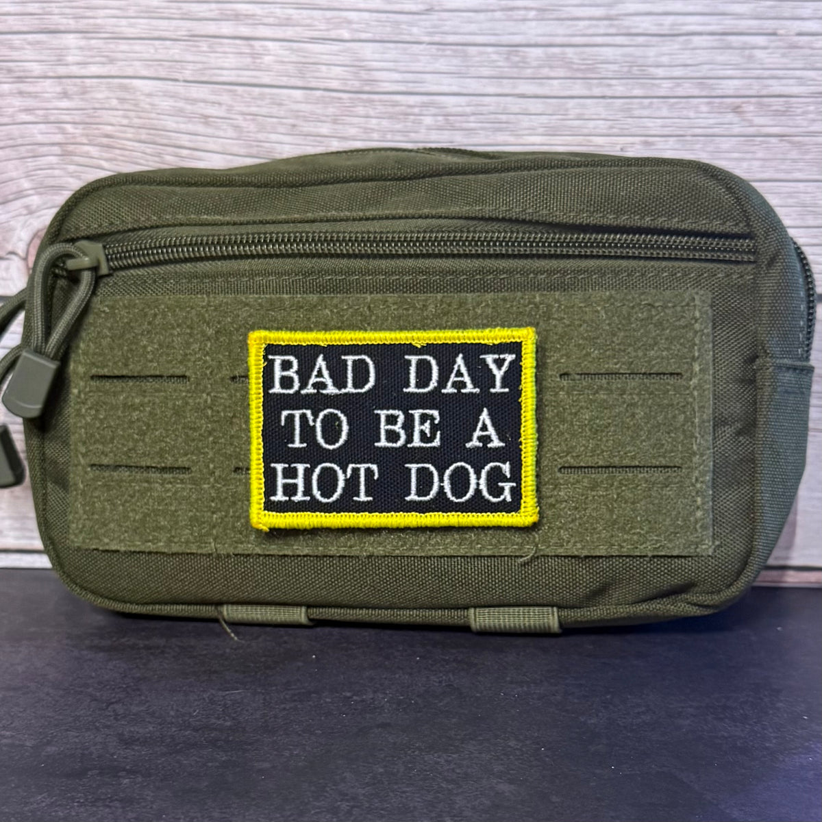 An olive green tactical pouch with a front zipper and a rectangular embroidered patch that reads 'BAD DAY TO BE A HOT DOG' in white letters on a black background with a yellow border, set against a light wood panel backdrop.