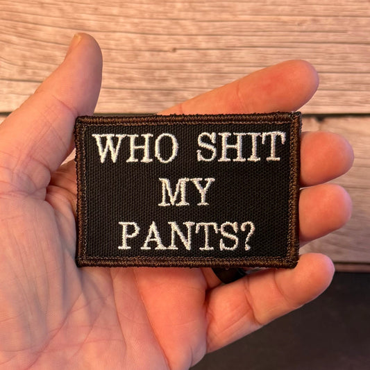 A hand holding a rectangular black embroidered patch with the text 'WHO SHIT MY PANTS?' in white capital letters, set against a wooden plank background.