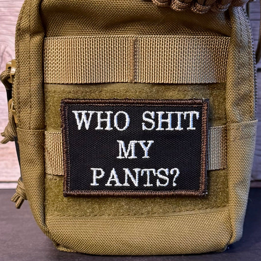 A close-up of a tactical pouch in olive drab and coyote brown, featuring a black rectangular patch with the white embroidered text 'WHO SHIT MY PANTS?'.