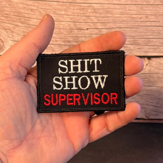 A hand holding a rectangular black embroidered patch that reads 'SHIT SHOW SUPERVISOR' in white and red lettering, with a wood plank background.