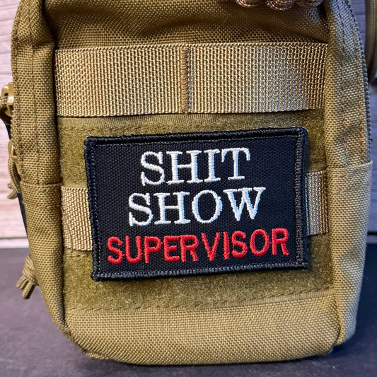 A close-up of a tactical pouch in olive drab nylon with a black embroidered patch reading 'SHIT SHOW SUPERVISOR' in white and red text. A brown paracord-wrapped strap is visible at the top, and the pouch is photographed against a rustic wood plank background.